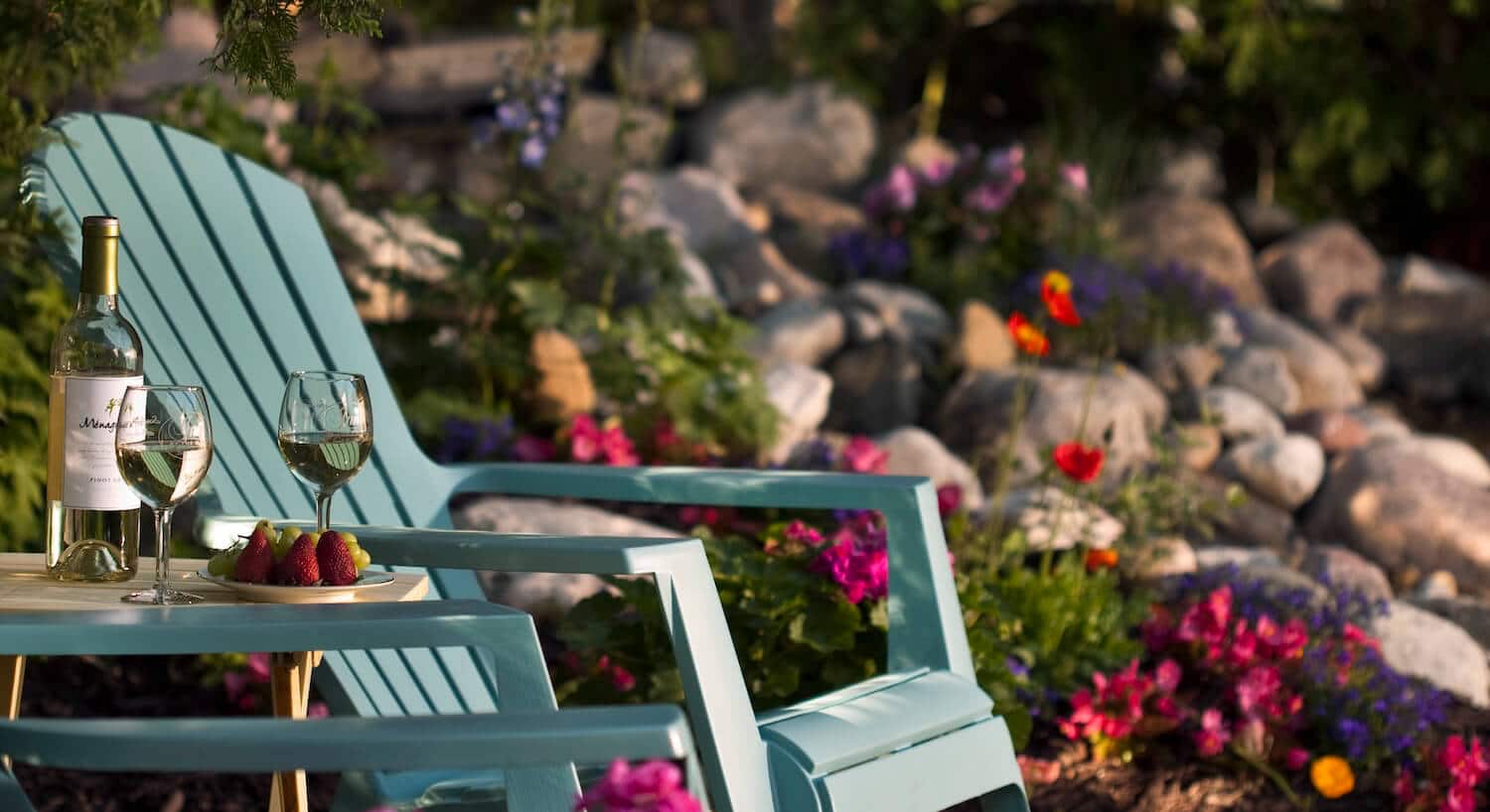 A tranquil outdoor setting features two turquoise chairs, a bottle of white wine, two glasses, and a plate of strawberries amid vibrant flowers and stones.