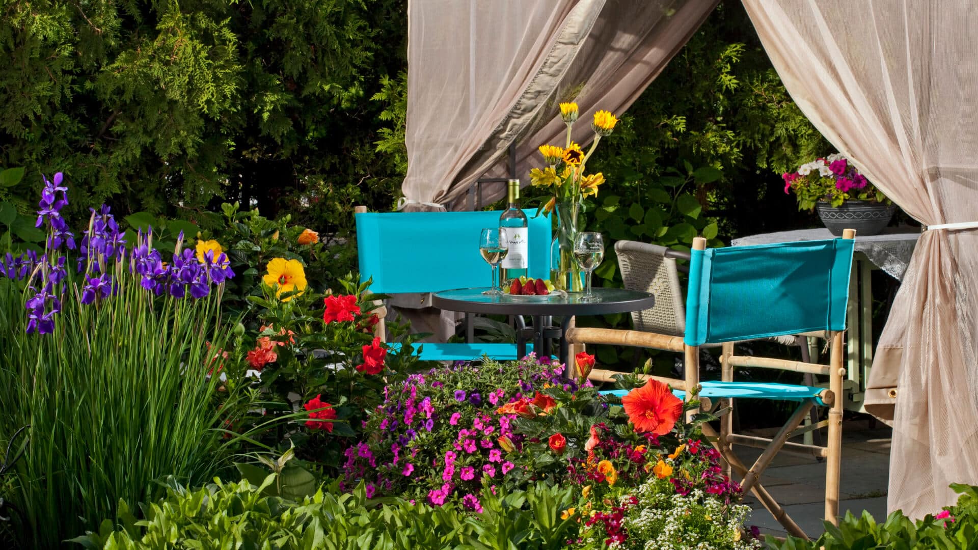 A cozy outdoor seating area surrounded by vibrant flowers and a bottle of wine on the table.