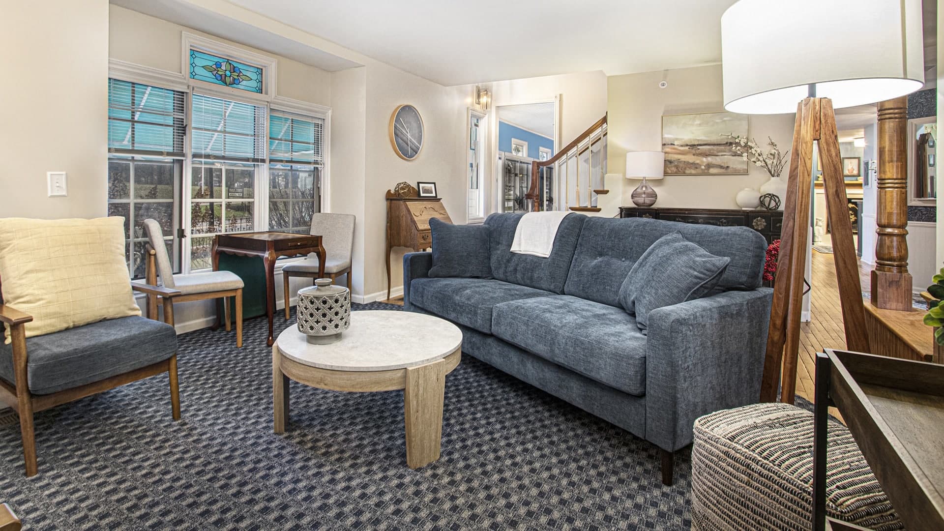 A cozy living room featuring a blue sofa, a round coffee table, and various seating options beside a window with decorative glass.