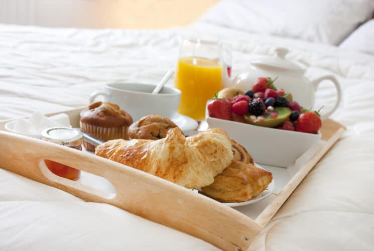 A breakfast tray with croissants, muffins, fruit, juice, and tea on a bed.