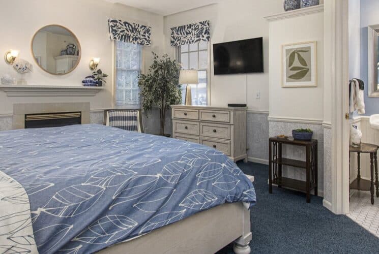 A bedroom with blue carpet, a wood framed bed with white and blue bedding, a blue sitting bench at the foot of the bed, a soaking tub built into a nook in the bedroom, and a sitting area with 2 plush chairs.