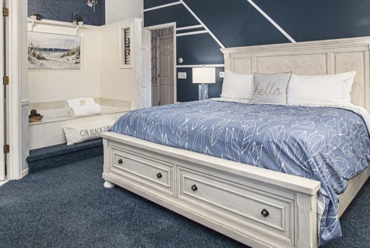 A bedroom with blue carpet, a wood framed bed with white and blue bedding, a blue sitting bench at the foot of the bed, a soaking tub built into a nook in the bedroom, and a sitting area with 2 plush chairs.