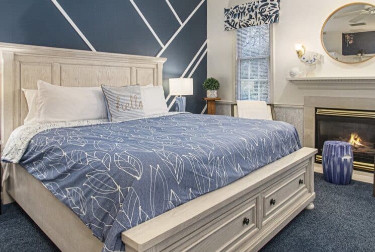 A bedroom with blue carpet, a wood framed bed with white and blue bedding, a blue sitting bench at the foot of the bed, a soaking tub built into a nook in the bedroom, and a sitting area with 2 plush chairs.